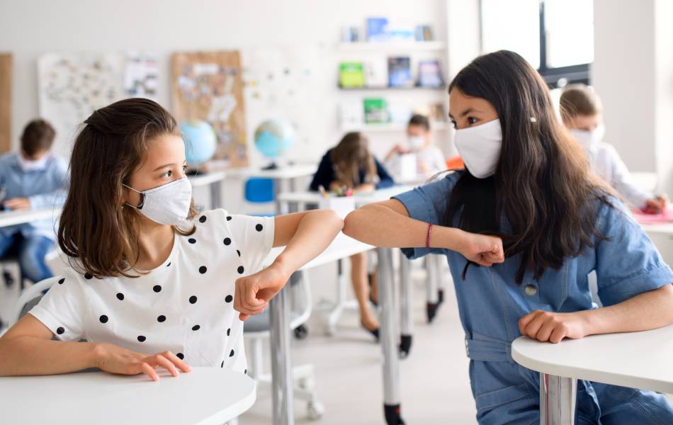Meninas dentro de uma sala de aula com máscaras se cumprimentando com o cotovelo devido ao covid-19