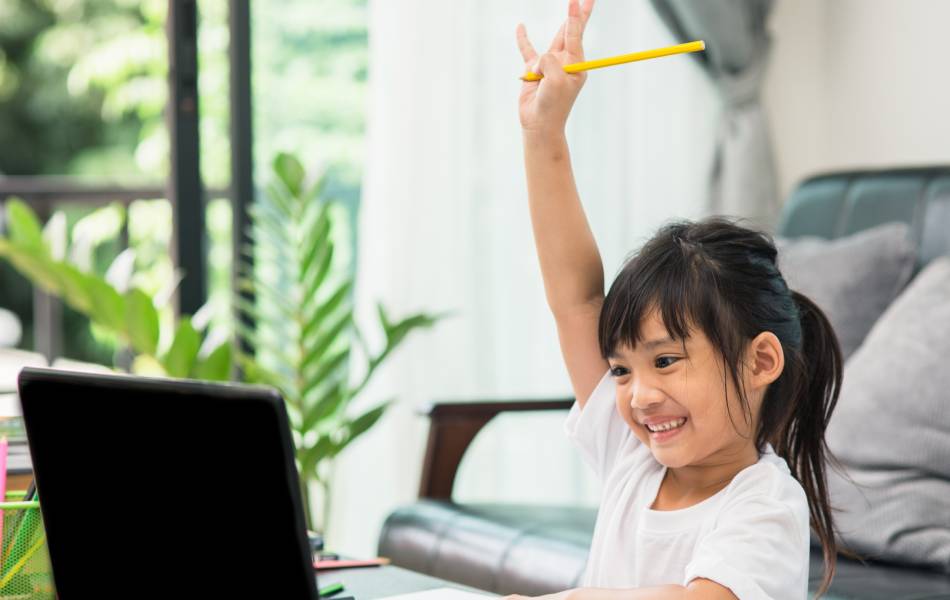 Menina fazendo aula ead em frente ao computador com a mão levantada, sinalizando que sabe a resposta da aula online