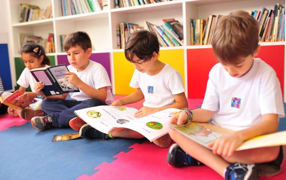 Crianças sentadas no chão com as pernas cruzadas estudando sobre educação socioemocional com um livro nas mãos
