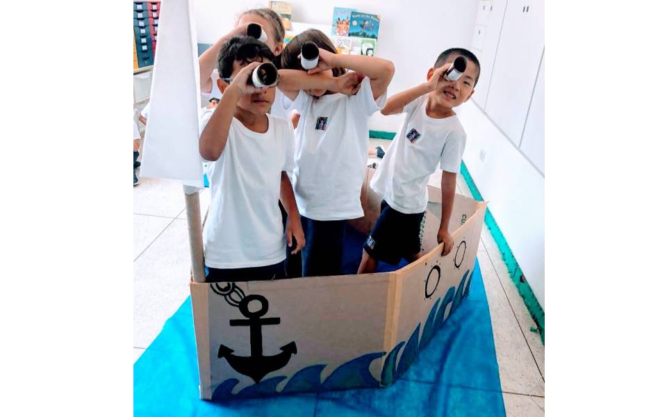 Crianças dentro de um barco feito em papelão simulando que estão navegando. Atividade de marinheiro em uma escola internacional