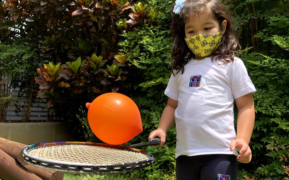 menina de máscara de proteção (covid-19) tentando equilibrar uma bexiga com uma raquete de tênis em uma escola bilíngue