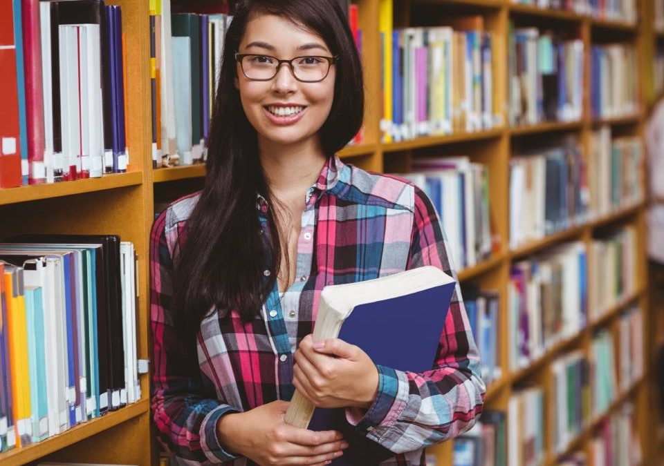 Mulher jovem dentro da biblioteca da escola ao lado de uma prateleira cheia de livros